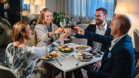Four individuals are enjoying a meal together at a restaurant. They are seated around a square table with dishes of food and are in the midst of a toast, with their wine glasses raised. The atmosphere appears convivial and celebratory.