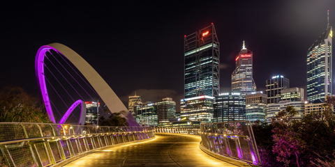 Night-time view of a pedestrian bridge in Perth. The bridge is lit up with footlights and tall buildings are lit up in the distance.